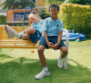 Duas crianças com uniformes azuis combinando, sentadas em um banco de praça.